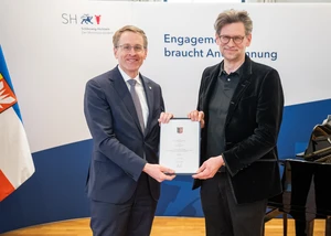 Zwei Männer mit Brille vor einer weißen Wand bei der Übergabe einer Urkunde