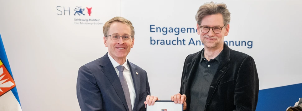 Zwei Männer mit Brille vor einer weißen Wand bei der Übergabe einer Urkunde