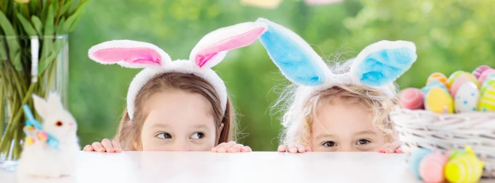 Mädchen mit Plüschhasenohren schauen auf eine weißen Korb mit bunten Ostereiern, © iStock.com/FamVeld