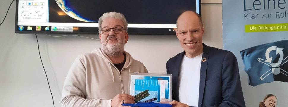 Zwei erwachsene Männer für einem Monitor und einer blauen Box in der Hand