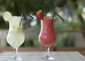 Zwei Cocktails, ein Mojito mit Limette und ein Erdbeer-Smoothie, auf einem Tisch in einer grünen Outdoor-Umgebung.