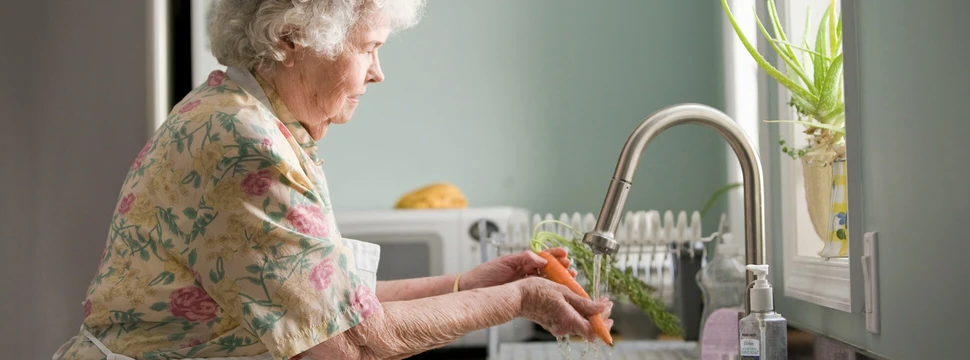 Eine Seniorin trägt ein geblümtes Kleid und wäscht in einer Küche Karotten