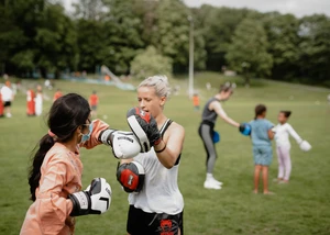 Zwei Frauen beim Boxtraining mit Handschuhen im Park, weitere Sportler im Hintergrund auf grüner Wiese.