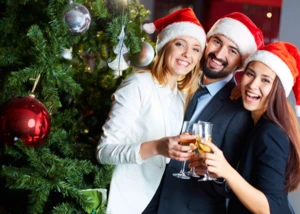 Weihnachtsfeier Drei Menschen mit Weihnachtsmützen neben einem geschmückten Tannenbaum