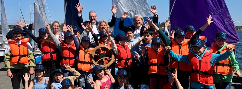 Kinder mit Schwimmwesten und Erwachsene stehen mit einem Steuerrad vor Segeln