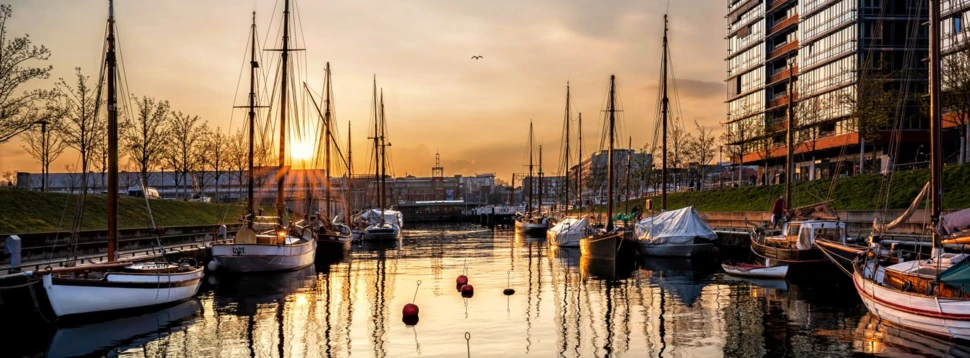 Sonnenuntergang über Segelbooten im modernen Stadthafen. Segelboote im Stadthafen bei Sonnenuntergang mit reflektierendem Wasser und moderner Architektur am Ufer.