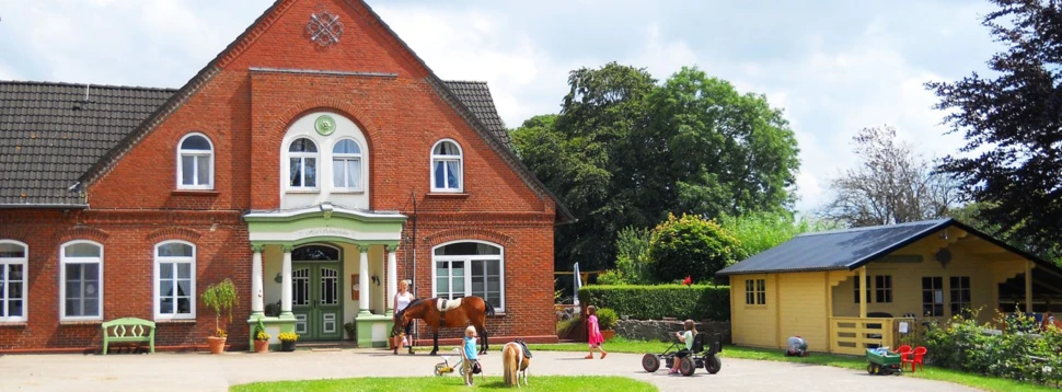Kinder und Tiere vor einem historischen Bauernhaus an einem Sommertag. Kinder spielen und reiten vor einem roten Backsteinhaus mit grüner Veranda.