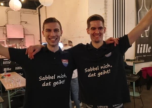 Zwei Männer in schwarzen T-Shirts, die gerade ein Darts Turnier gewonnen haben