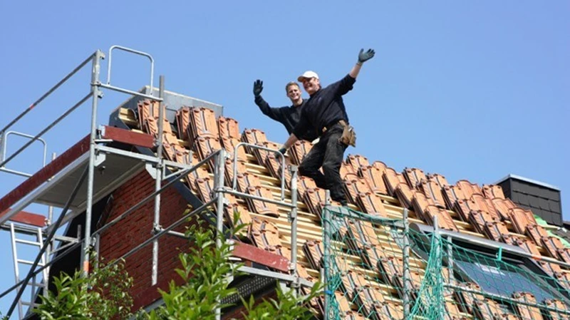 Zwei Mitarbeiter winken fröhlich vom Gerüst eines zu sanierenden Hauses mit Tonziegeln bei blauem Himmel.