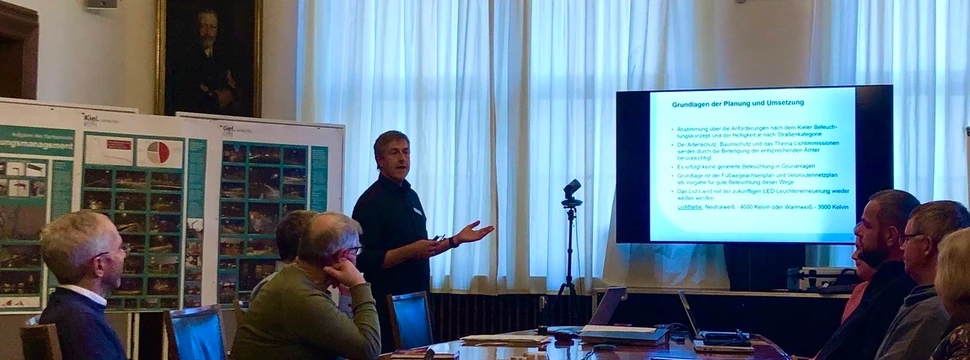 Präsentation in elegantem Sitzungssaal mit Kronleuchtern, Referent vor Bildschirm und Zuhörern am Konferenztisch