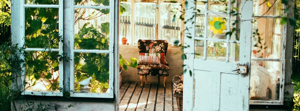 Ein offenes Gartentor führt von ihnen auf eine sonnige Veranda mit Pflanzen und Stuhl