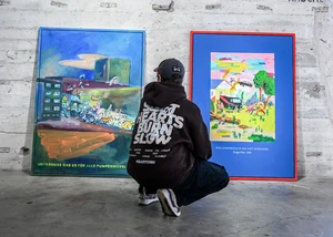 Ein Mann in einem schwarzen Pullover mit einer schwarzen Cap vor einer Wand, an der zwei Bilder lehnen