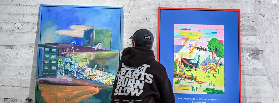 Ein Mann in einem schwarzen Pullover mit einer schwarzen Cap vor einer Wand, an der zwei Bilder lehnen