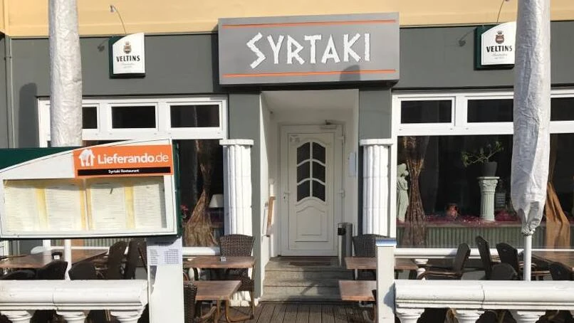 Das Restaurant Syrtaki Kiel von der Straße aus Eine Veranda mit Sonnenschirmen, einer Tür und einem Schild auf dem "Syrtaki" steht