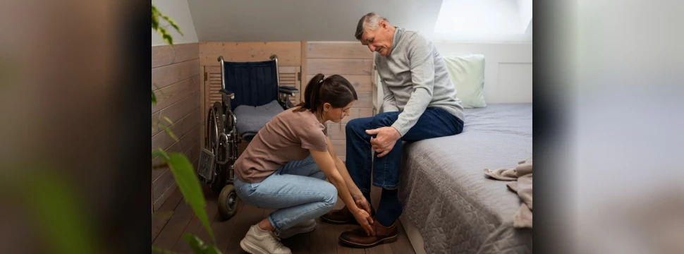 Alltägliche Pflege mit Herz im eigenen Zuhause. Junge Frau hilft älterem Mann beim Anziehen der Schuhe neben Rollstuhl im Schlafzimmer.