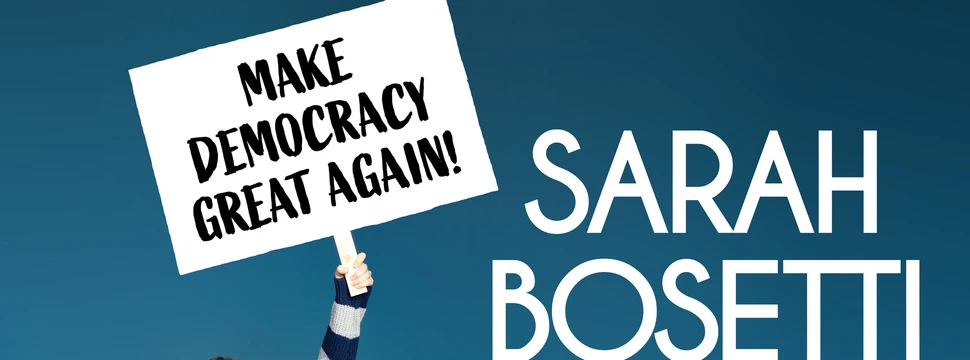 Frau sitzt auf einem Sofa und hält ein Schild hoch mit der Aufschrift: Make democracy great again