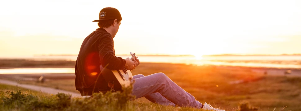 Ein Mann mit einer umgedrehten Cap auf einem Deich mit einer Gitarre in seiner Hand und dem Sonnenuntergang im Hintergrund
