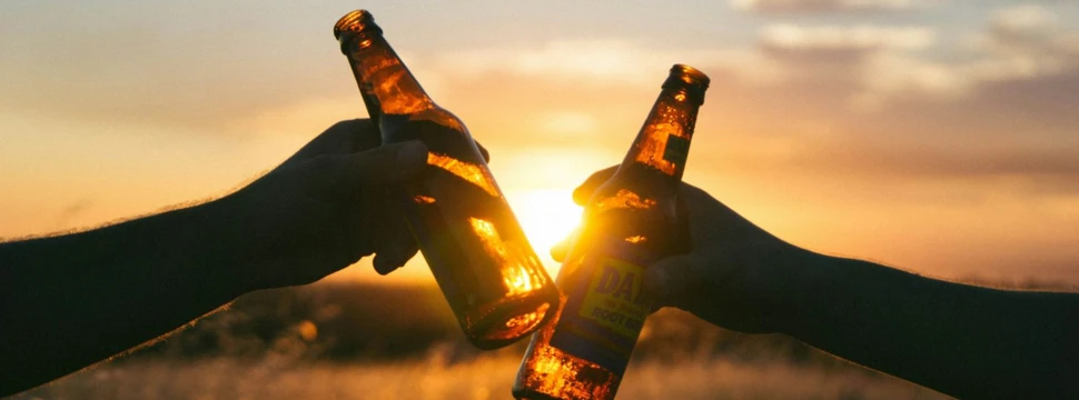 Zwei Bierflaschen werden aneinandergestoßen, im Hintergrund Sonnenuntergang
