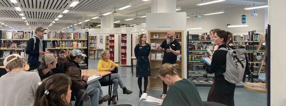 Mehrere Menschen in einem Raum mit Deckenlampen und Regalen im Hintergrund, die sich beraten und austauschen