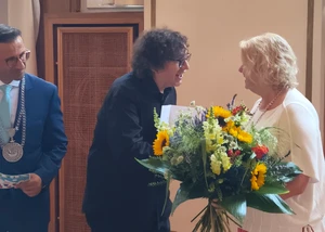 Ein Mann in einem dunklen Anzug zwischen zwei anderen Menschen, die gerade einen Blumenstrauß überreichen