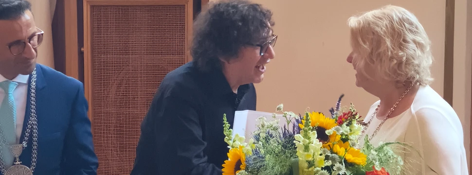 Ein Mann in einem dunklen Anzug zwischen zwei anderen Menschen, die gerade einen Blumenstrauß überreichen