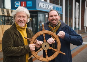 Zwei Männer vor einem Gebäude, die ein Schiffsteuerrad in der Hand haben