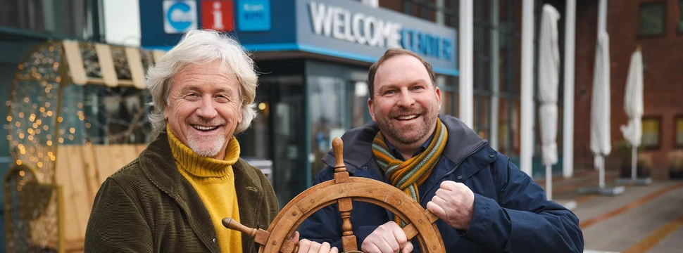 Zwei Männer vor einem Gebäude, die ein Schiffsteuerrad in der Hand haben