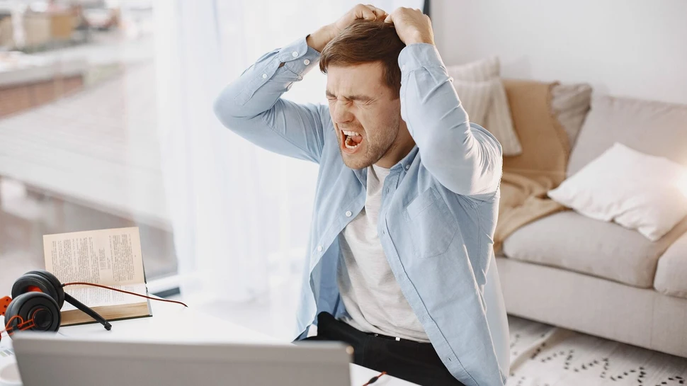 Frustrierter junger Mann im blauen Hemd greift sich schreiend mit beiden Händen in die Haare, während er vor einem Laptop am Schreibtisch sitzt.