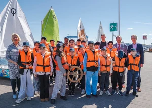 Mehrere Menschen an der Kieler Förde, davon viele in orangenen Rettungswesten, an der Kiellinie vor mehreren Segelbooten