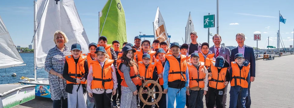 Mehrere Menschen an der Kieler Förde, davon viele in orangenen Rettungswesten, an der Kiellinie vor mehreren Segelbooten