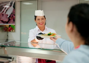 Eine Frau in einem weißen Shirt, die auch eine weiße Kochmütze und schwarze Handschuhe trägt, übergibt einen Teller mit Essen an eine andere Person