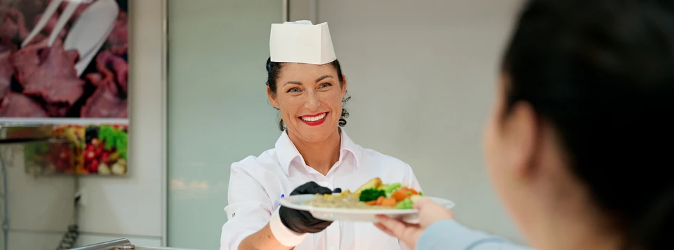 Eine Frau in einem weißen Shirt, die auch eine weiße Kochmütze und schwarze Handschuhe trägt, übergibt einen Teller mit Essen an eine andere Person