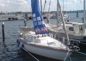 Weiße Segelyacht mit blauer Flagge im Hafen, umgeben von anderen Booten und Stegen bei sonnigem Wetter.