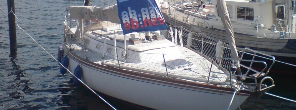 Weiße Segelyacht mit blauer Flagge im Hafen, umgeben von anderen Booten und Stegen bei sonnigem Wetter.