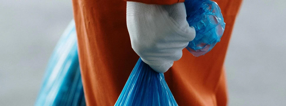 Ein blauer Müllsack in der Hand eines Menschens, der einen weißen Handschuh und eine orangene Hose trägt