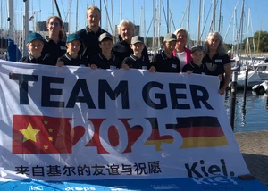 Die Coaches Alexa und Noah, GF Uwe Wanger, Stadtpräsidentin Bettina Aust, Pressesprecherin Eva Zeiske und im Vordergrund die sechs auserwählten Segelkinder Eine Gruppe von Menschen mit einem Banner in der Hand, der auf die Partnerschaft zwischen Kiel und der Stadt Qingdao hinweist