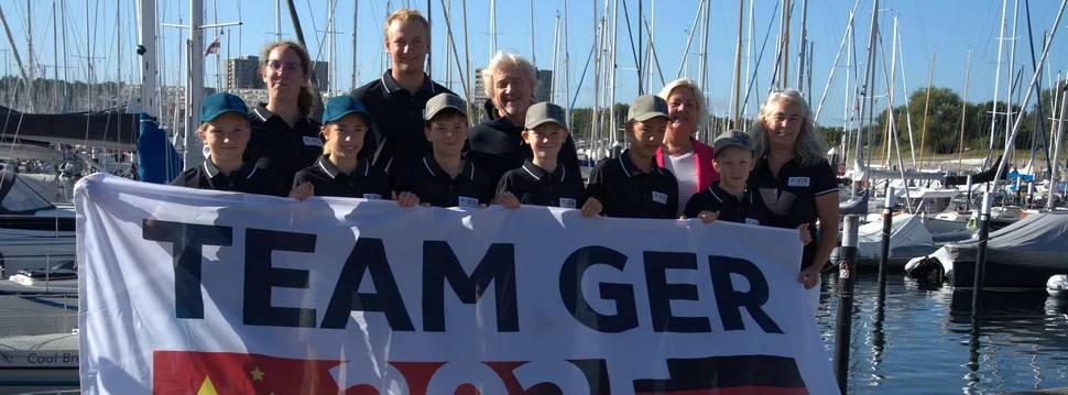 Die Coaches Alexa und Noah, GF Uwe Wanger, Stadtpräsidentin Bettina Aust, Pressesprecherin Eva Zeiske und im Vordergrund die sechs auserwählten Segelkinder Eine Gruppe von Menschen mit einem Banner in der Hand, der auf die Partnerschaft zwischen Kiel und der Stadt Qingdao hinweist