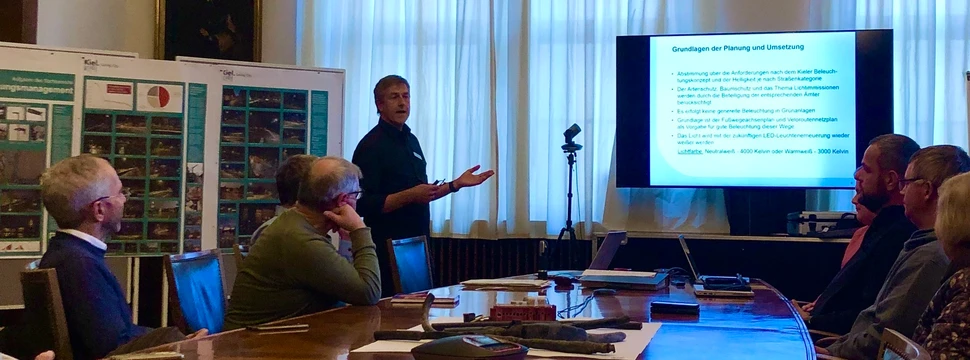 Eine Fachpräsentation in einem klassischen Rathaus-Sitzungssaal. Präsentation in elegantem Sitzungssaal mit Kronleuchtern, Referent vor Bildschirm und Zuhörern am Konferenztisch.