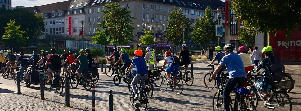 Radfahrende in einer Gruppe auf einem sonnigen Platz mit historischen Gebäuden im Hintergrund.