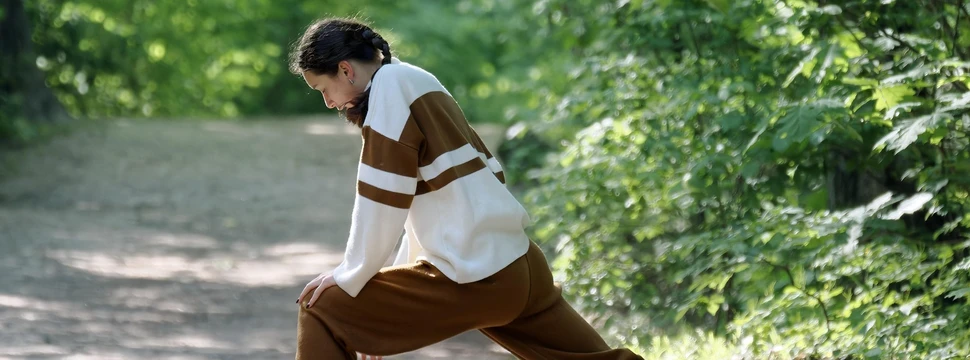 Eine Frau in braun-weißer Sportkleidung, die sich vor grünen Pflanzen am Dehnen ist