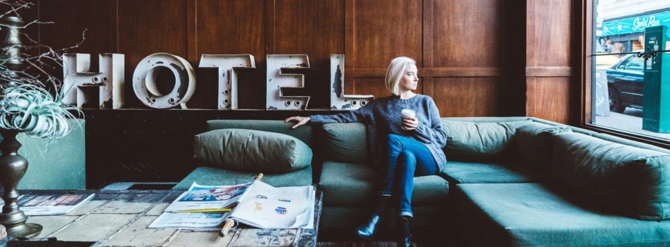 Entspannter Moment in einer stilvollen Hotellobby mit Blick auf die Stadt. Frau sitzt mit Kaffee in moderner Hotellobby auf Couch und blickt aus dem Fenster auf die Stadt.