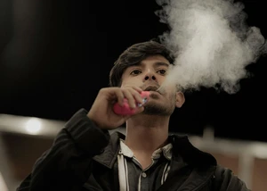 Junger Mann in dunkler Jacke sitzt in einem abgedunkelten Raum und atmet beim Vapen mit einer pinken E-Zigarette eine große weiße Dampfwolke aus.
