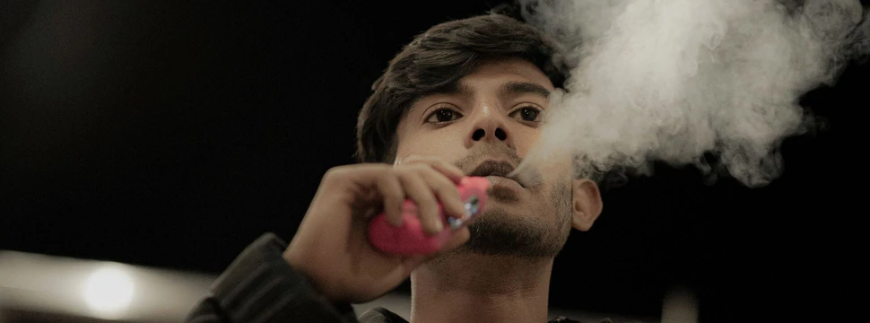 Junger Mann in dunkler Jacke sitzt in einem abgedunkelten Raum und atmet beim Vapen mit einer pinken E-Zigarette eine große weiße Dampfwolke aus.