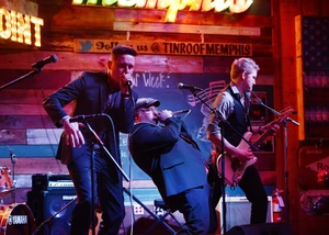 Drei Menschen auf einer Bühne während eines Auftritts mit Mikrophonen im Vordergrund und mit Instrumenten in der Hand