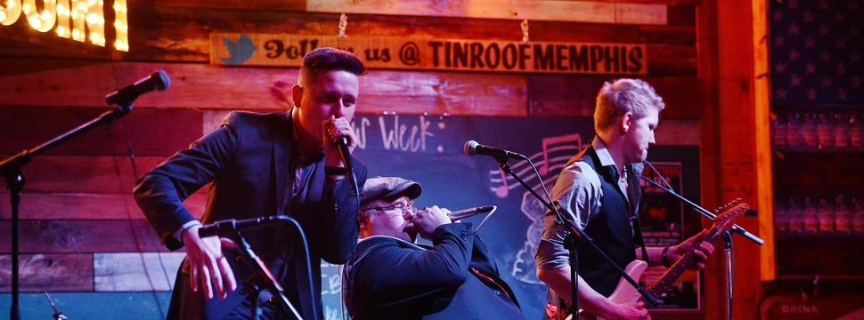 Drei Menschen auf einer Bühne während eines Auftritts mit Mikrophonen im Vordergrund und mit Instrumenten in der Hand