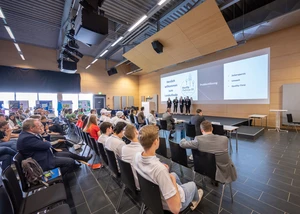 Mehrere Menschen auf Stühlen sitzend vor einer Bühne, auf der eine Präsentation stattfindet