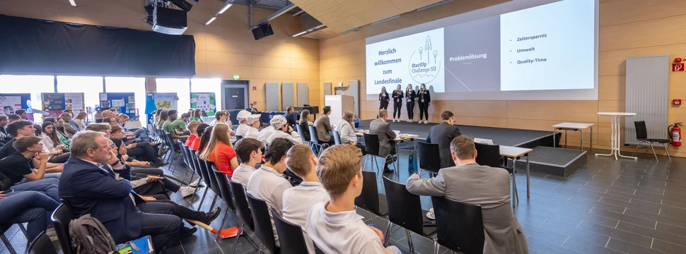 Mehrere Menschen auf Stühlen sitzend vor einer Bühne, auf der eine Präsentation stattfindet