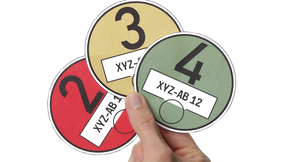 Eine Hand hält drei Umweltplaketten in unterschiedlichen Farben – rot, gelb und grün.