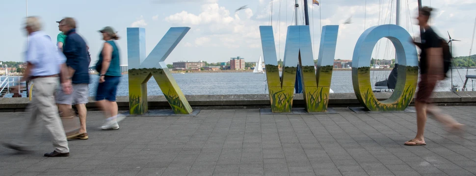 Große Lettern an einem Hafen, davor Menschen