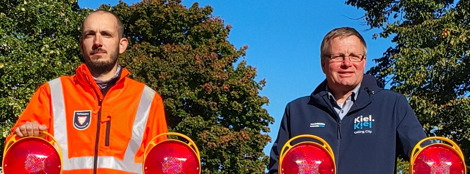 Zwei Männer, davon einer in einer orangenen Weste mit der Aufschrift "Tiefbauamt" und der andere in einer dunkelblauen mit der Aufschrift "Kiel", vor einer Schranke im Kieler Stadtteil Mettenhof
